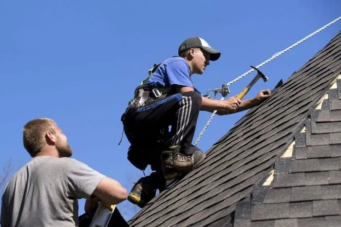 A roofer in a fall-protection harness nailing asphalt shingles